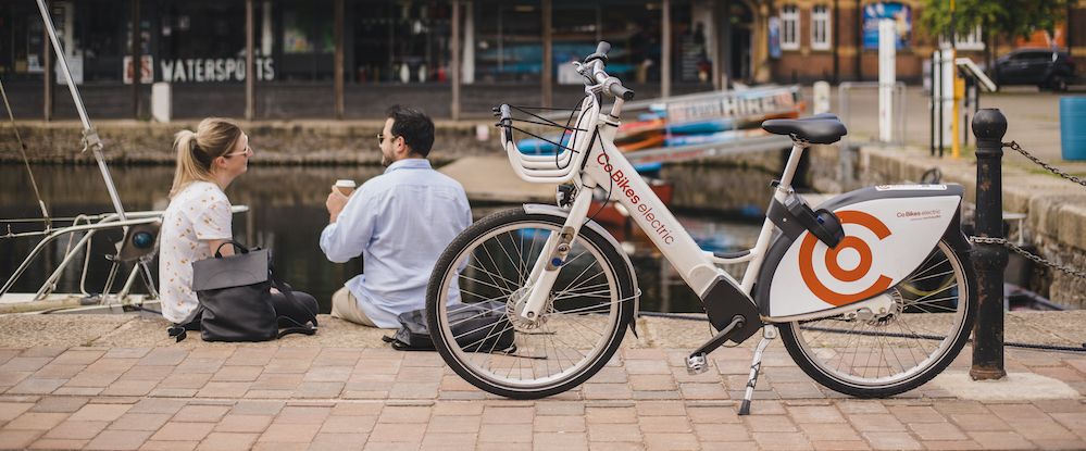 Photo of a Co Bikes bike in the foregorund, with a women and man sat on Exeter Quayside in the background